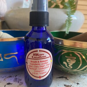 May include: A blue glass bottle with a black spray top and a white label that reads "RHaw Creations" and "Breath & Release". The label lists the ingredients: purified water, Clary Sage, Dark Patchouli, Cedarwood, Cinnamon. The bottle is 2 fl oz and is labeled "organic".