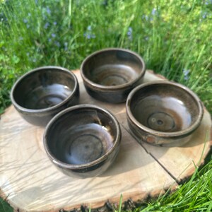 May include: Four handmade ceramic bowls with a dark, earthy glaze. The bowls are arranged on a natural wood slice, showcasing their rustic charm. The bowls are small, perfect for serving dips or snacks. The background features green grass and wildflowers.