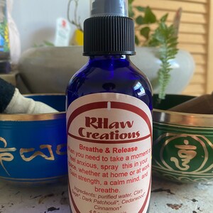 May include: A blue glass bottle with a black spray nozzle and a white label that reads "RHaw Creations" and "Breathe & Release". The label also lists the ingredients: purified water, Clary Sage, Dark Patchouli, Cedarwood, Cinnamon. The bottle contains 4 fl oz. of organic essential oil blend.