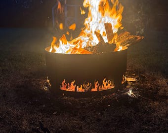 May include: A round, dark metal fire pit with flame cutouts around the base. Bright orange and yellow flames rise from burning wood inside the pit. The fire pit is set on a grassy area at night.