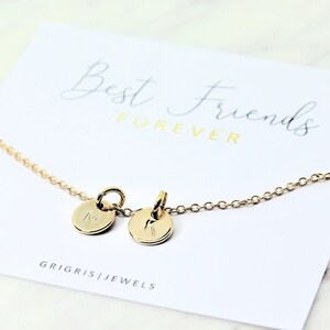 May include: A gold-colored necklace with two small, round pendants, each engraved with a letter. The necklace is displayed on a white card with the words "Best Friends Forever" and the brand name "GRIGRIS | JEWELS".