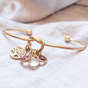 May include: Gold bangle bracelet with a knot design and multiple charms, including a tree of life charm and several small gemstone charms.