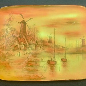 May include: Rectangular serving tray with a painted landscape scene. The artwork depicts windmills, houses, and boats on a body of water, set against a warm, sunset-colored sky. The tray has rounded corners and a gold-colored trim.