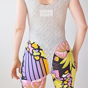 Pode incluir: Um body de manga curta com uma secção superior cinzenta clara texturada e uma pequena etiqueta retangular branca com o texto C&L. A secção inferior apresenta uma estampa gráfica multicolorida vibrante com motivos florais amarelos, roxos e pretos.