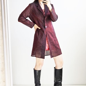 Pode incluir: Um blazer de manga comprida e comprimento até ao joelho com um padrão manchado em bordeaux escuro e preto. O blazer apresenta lapelas entalhadas, fecho frontal com dois botões e bainha reta. Combinado com botas altas de couro preto.