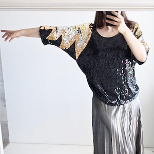 Op de afbeelding: Een zwart topje met lovertjes en goud- en zilverkleurige lovertjes op de schouders, gecombineerd met een metallic zilveren geplooide midirok en zwarte laarzen met een spitse neus. De top heeft korte mouwen en een ronde halslijn.