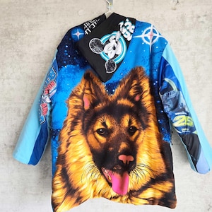 Pu&ograve; includere: Una giacca a maniche lunghe con una grande e dettagliata grafica di un cane pastore tedesco sul retro. La giacca presenta uno sfondo blu vivace con motivi a stelle, accenti neri e ulteriori testi grafici e loghi sulle maniche e sul cappuccio.