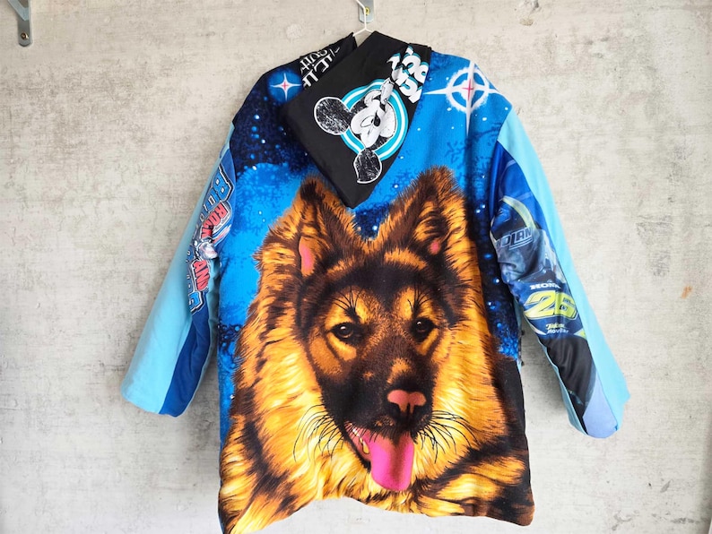 Pu&ograve; includere: Una giacca a maniche lunghe con una grande e dettagliata grafica di un cane pastore tedesco sul retro. La giacca presenta uno sfondo blu vivace con motivi a stelle, accenti neri e ulteriori testi grafici e loghi sulle maniche e sul cappuccio.
