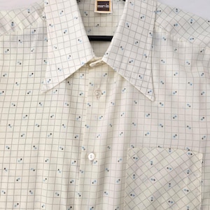 Può includere: Camicia color crema con bottoni e un motivo a griglia di piccoli quadrati neri e blu. La camicia ha un colletto a punta, una tasca sul petto e il marchio "marvin" su un'etichetta vicino al colletto.