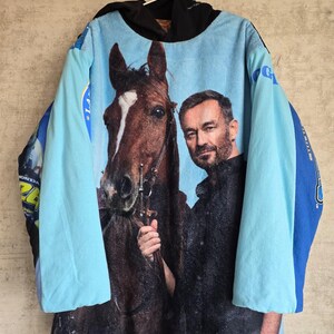 Pu&ograve; includere: Una felpa con cappuccio a maniche lunghe caratterizzata da una grande stampa fotografica di un cavallo marrone e una persona barbuta sul davanti. Il capo presenta maniche azzurre, un cappuccio nero e pannelli laterali blu.