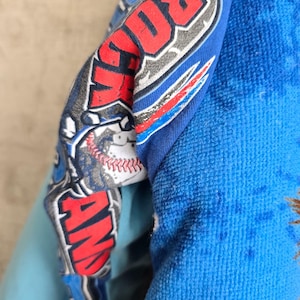 Pu&ograve; includere: Un capo in tessuto blu con una stampa grafica che riporta il testo ROCK AND, un'illustrazione di una palla da baseball con cuciture rosse e accenti stilizzati blu e rossi. Il materiale ha una superficie morbida e testurizzata con pieghe.