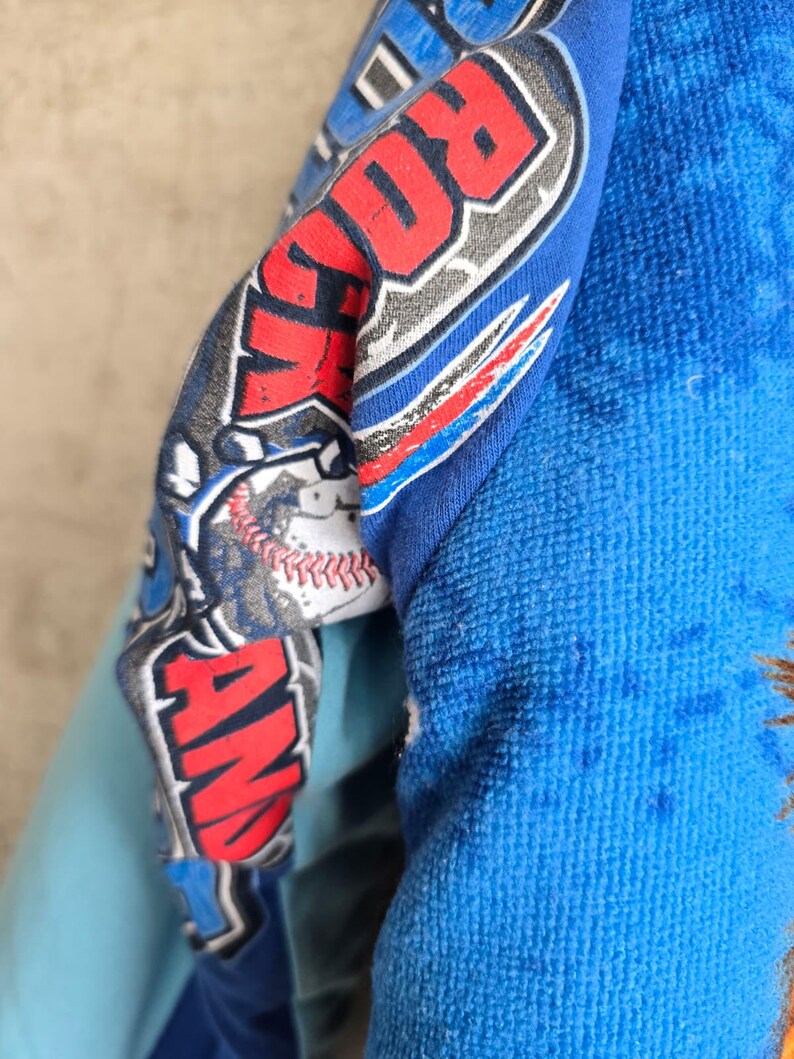 Pu&ograve; includere: Un capo in tessuto blu con una stampa grafica che riporta il testo ROCK AND, un'illustrazione di una palla da baseball con cuciture rosse e accenti stilizzati blu e rossi. Il materiale ha una superficie morbida e testurizzata con pieghe.