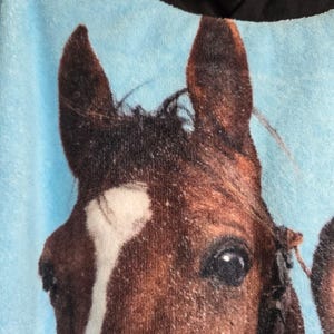 Pu&ograve; includere: Un morbido capo con cappuccio azzurro caratterizzato da una grande stampa fotografica dettagliata della testa di un cavallo marrone. Il cavallo presenta una vistosa macchia bianca sulla fronte e occhi scuri. Il cappuccio &egrave; di colore nero.