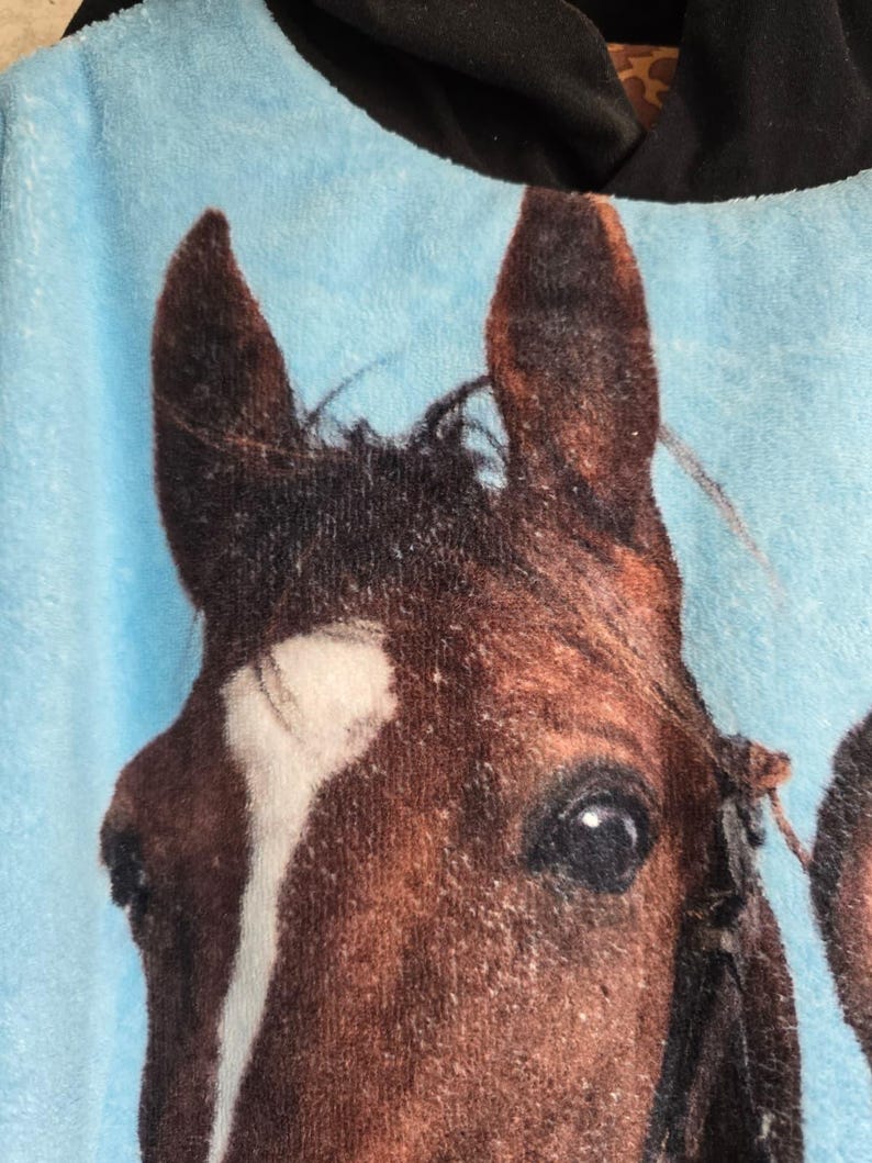 Pu&ograve; includere: Un morbido capo con cappuccio azzurro caratterizzato da una grande stampa fotografica dettagliata della testa di un cavallo marrone. Il cavallo presenta una vistosa macchia bianca sulla fronte e occhi scuri. Il cappuccio &egrave; di colore nero.