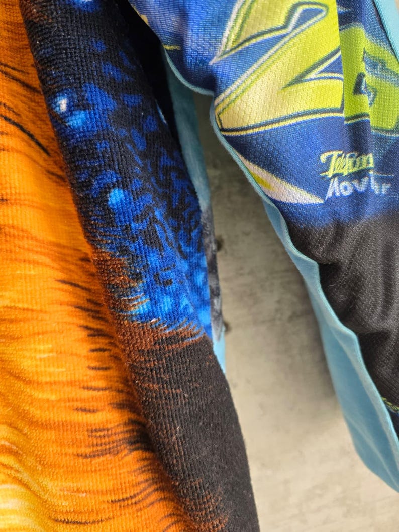 Pu&ograve; includere: Un capo in tessuto testurizzato caratterizzato da una sezione arancione vivace con linee orizzontali scure, una sezione blu scuro con motivo maculato e una manica azzurra che mostra il testo giallo "26" e la parola "Movistar" in bianco.