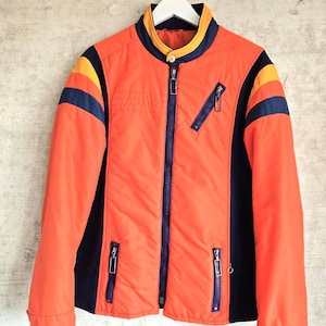 May include: A bright orange jacket featuring a mandarin collar with yellow and navy blue trim. The jacket includes navy blue side panels, navy blue stripes on the shoulders, and three zippered pockets with navy blue accents and silver hardware.