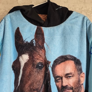 Pu&ograve; includere: Una felpa con cappuccio azzurra caratterizzata da una grande stampa grafica di un cavallo marrone con una macchia bianca sul muso accanto al ritratto di un uomo con la barba. Il capo presenta un cappuccio nero e una consistenza morbida e felpata.