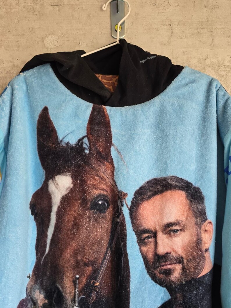 Pu&ograve; includere: Una felpa con cappuccio azzurra caratterizzata da una grande stampa grafica di un cavallo marrone con una macchia bianca sul muso accanto al ritratto di un uomo con la barba. Il capo presenta un cappuccio nero e una consistenza morbida e felpata.