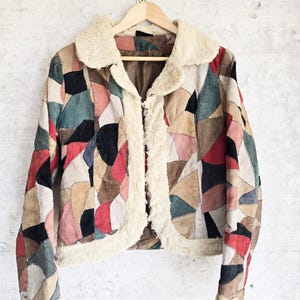 Könnte beinhalten: Eine Patchwork-Jacke mit mehrfarbigen Wildledereinsätzen in Rot, Schwarz, Beige und Blaugrün. Die Jacke verfügt über einen cremefarbenen Kunstfellkragen, eine vordere Zierleiste und Ärmelbündchen sowie ein braunes Innenfutter am Halsausschnitt.