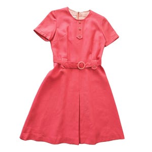 Puede incluir: Un vestido vintage de color coral con mangas cortas y corpiño ajustado. El vestido presenta un cinturón a juego con una hebilla circular y una falda plisada. El escote tiene un pequeño cuello puntiagudo y tres botones decorativos.