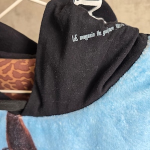 Pu&ograve; includere: Una felpa con cappuccio nera che riporta la scritta bianca LE magasin de guitare, un corpo principale in tessuto azzurro soffice e un motivo animalier marrone e nero visibile sulla fodera interna del cappuccio, appesa a una gruccia bianca.