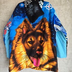 Pu&ograve; includere: Una maglia a maniche lunghe con una grande e dettagliata grafica di un pastore tedesco con la lingua rosa brillante. Lo sfondo &egrave; una vibrante scena spaziale blu con stelle ed elementi testuali. &Egrave; attaccato uno scaldacollo nero con testo bianco.