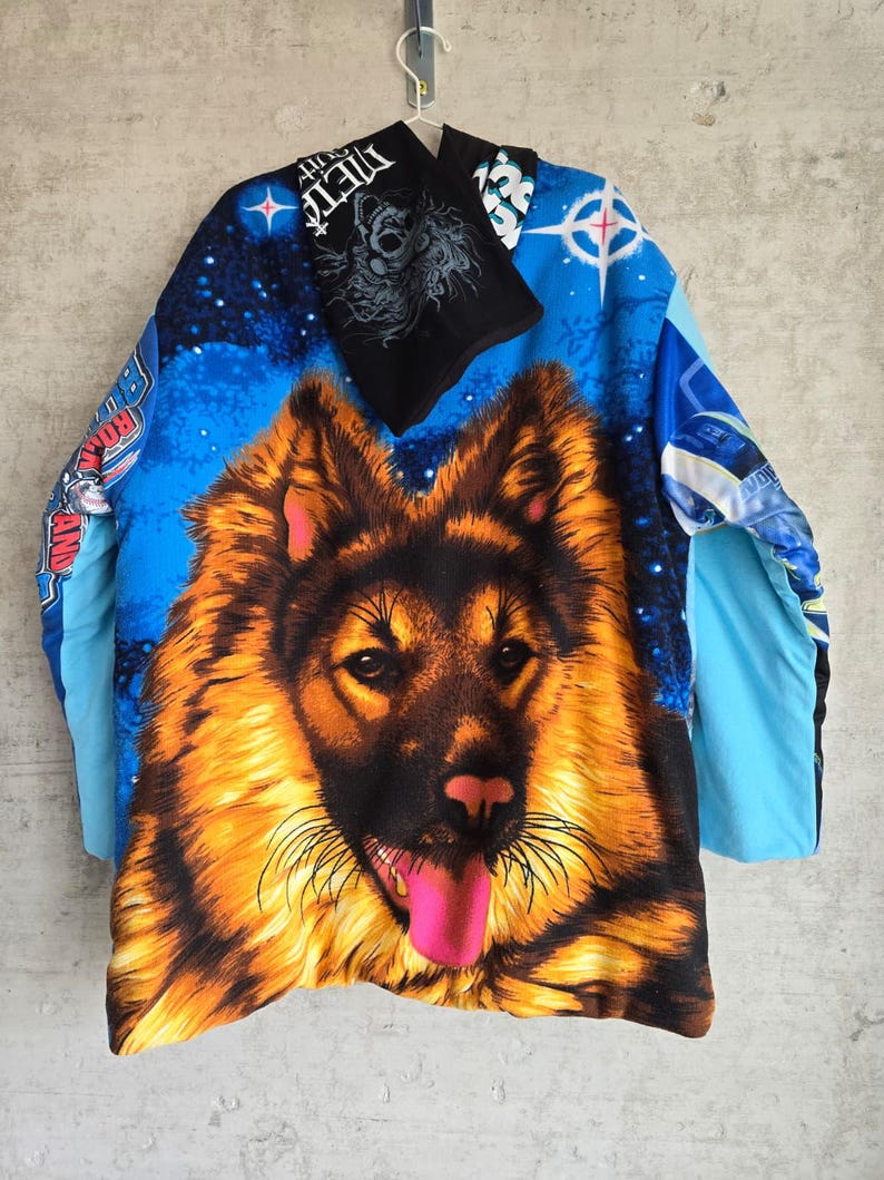 Pu&ograve; includere: Una maglia a maniche lunghe con una grande e dettagliata grafica di un pastore tedesco con la lingua rosa brillante. Lo sfondo &egrave; una vibrante scena spaziale blu con stelle ed elementi testuali. &Egrave; attaccato uno scaldacollo nero con testo bianco.