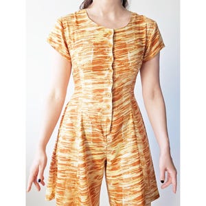 Op de afbeelding: Vintage jumpsuit in oranje en gele tinten. Het kledingstuk met korte mouwen heeft een knoopsluiting aan de voorkant, een getailleerd lijfje en een korte broek. De stof heeft een gestructureerd, abstract patroon. Klassieke retro-stijl.