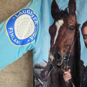 Pu&ograve; includere: Una felpa con cappuccio azzurra caratterizzata da una grande stampa fotografica di un cavallo marrone e una persona. La manica mostra un logo circolare con il testo LAUGHTER YOGA FINLAND e un motivo a spirale della parola HA ripetuta.