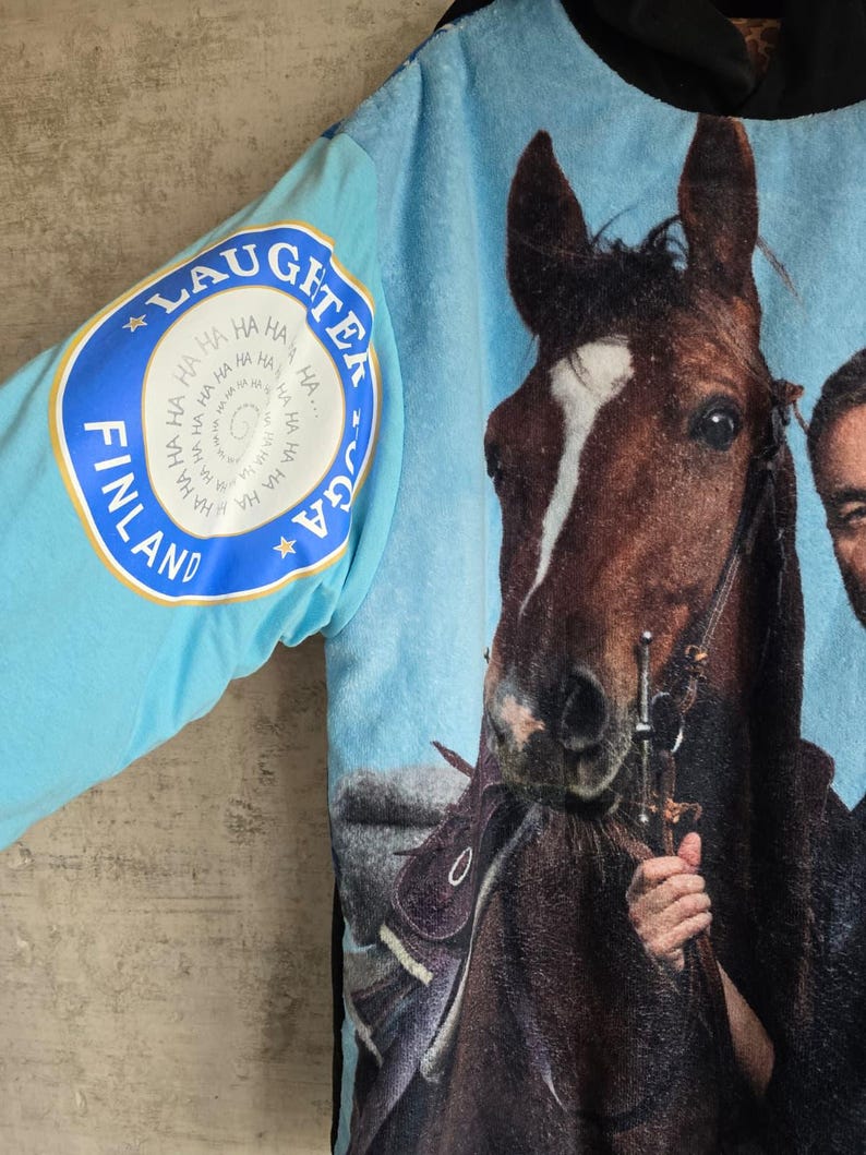 Pu&ograve; includere: Una felpa con cappuccio azzurra caratterizzata da una grande stampa fotografica di un cavallo marrone e una persona. La manica mostra un logo circolare con il testo LAUGHTER YOGA FINLAND e un motivo a spirale della parola HA ripetuta.