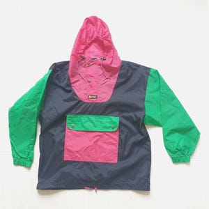 Könnte beinhalten: Eine marineblaue Schlupfjacke mit einer leuchtend pinken Kapuze und einer passenden pinken Vordertasche mit grünem Umschlag. Die Ärmel sind grün, und die Jacke verfügt über eine Kapuze mit Kordelzug, eine Teilknopfleiste und ein kleines Logo-Patch.