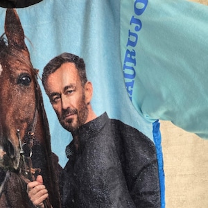 Pu&ograve; includere: Una felpa con cappuccio azzurra con una grande stampa grafica di una persona con un cavallo marrone. La persona indossa una camicia nera. La manica presenta la scritta AURUJOO in blu. Cappuccio nero e pannello laterale blu testurizzato.
