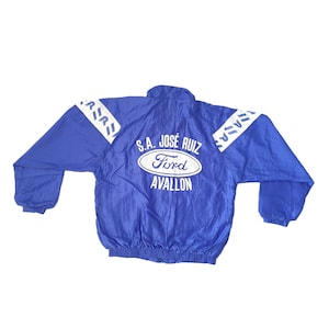 Op de afbeelding: Blauw nylon jack met een witte rits en witte bies. Het jack heeft de tekst "S.A. JOSE RUIZ Ford AVALLON" op de achterkant.