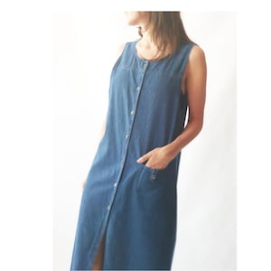 May include: Sleeveless, button-down denim dress in a medium blue wash. The dress features a round neckline, two front pockets, and a knee-length hemline. The dress is made of a soft denim fabric and has a relaxed fit.