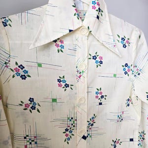 Puede incluir: Camisa crema con botones y cuello grande, adornada con un estampado floral en rosa, azul y verde. La camisa presenta un diseño de cuadrícula geométrica con líneas finas. La camisa está colgada en una percha blanca.