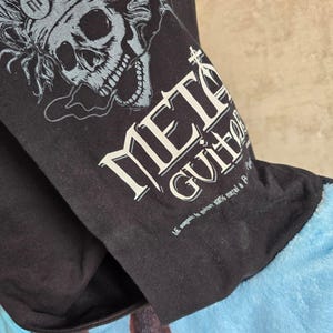 Pu&ograve; includere: Una t-shirt nera con la grafica di un teschio che indossa cuffie e il testo "METAL GUITAR" in un carattere audace e stilizzato. Sotto il testo principale, una scritta bianca pi&ugrave; piccola recita "LE magasin de guitare 100% metal &agrave; Paris".