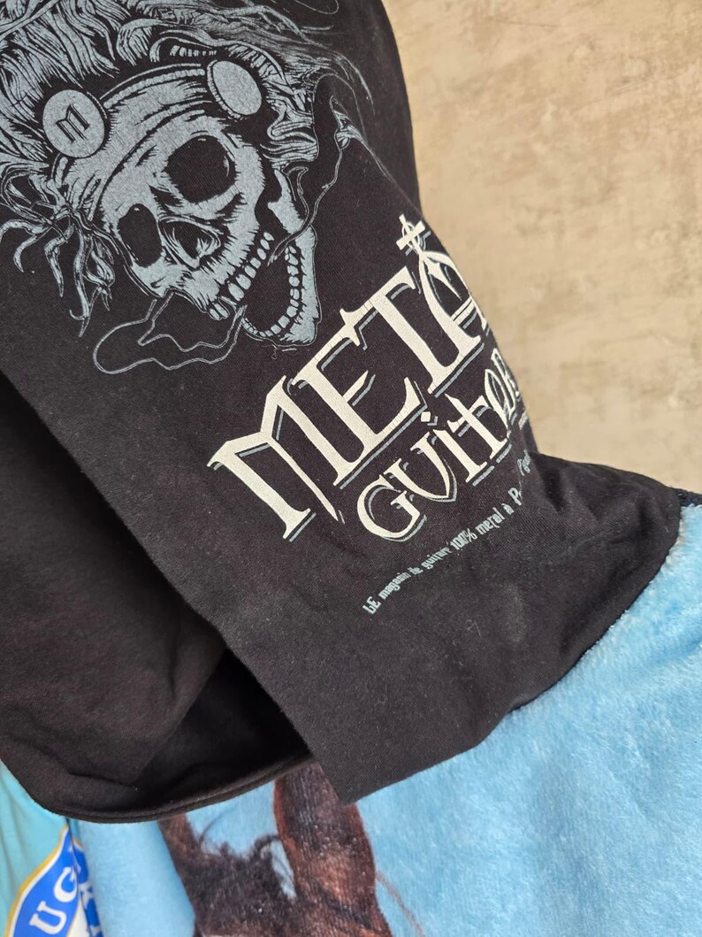 Pu&ograve; includere: Una t-shirt nera con la grafica di un teschio che indossa cuffie e il testo "METAL GUITAR" in un carattere audace e stilizzato. Sotto il testo principale, una scritta bianca pi&ugrave; piccola recita "LE magasin de guitare 100% metal &agrave; Paris".