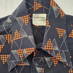 Puede incluir: Una camisa negra de botones con un patrón geométrico de triángulos de pata de gallo naranja y líneas punteadas blancas. La etiqueta del cuello muestra el texto pointer, finest Finnish quality y 100% cotton con símbolos de cuidado.
