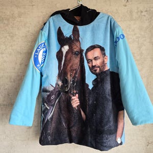 Pu&ograve; includere: Una felpa con cappuccio azzurra con una grande stampa grafica di un cavallo marrone e un uomo sul davanti. Le maniche sono azzurre e il cappuccio &egrave; nero. Un emblema circolare blu con testo bianco &egrave; visibile sulla manica destra.