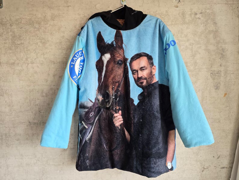 Pu&ograve; includere: Una felpa con cappuccio azzurra con una grande stampa grafica di un cavallo marrone e un uomo sul davanti. Le maniche sono azzurre e il cappuccio &egrave; nero. Un emblema circolare blu con testo bianco &egrave; visibile sulla manica destra.