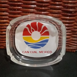 Puede incluir: Cenicero de vidrio transparente, de forma rectangular con esquinas redondeadas. El cenicero presenta un gráfico colorido de un sol sobre el océano, con el texto "CAN CUN, MEXICO" impreso debajo. El cenicero está sobre una superficie negra.