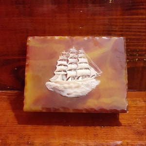 May include: A rectangular, amber-colored box with a carved white sailing ship design. The box has a scalloped edge and sits on a wooden surface. The ship has three masts and is depicted on a sea of white.