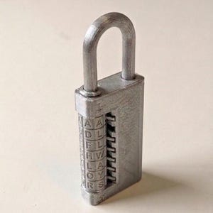 Può includere: Un lucchetto a combinazione argentato con un arco curvo. La serratura presenta una serie di quadranti rotanti con lettere. Le lettere visibili sui quadranti sono A, D, F, H, L, O e R. Il lucchetto è realizzato in un materiale metallico strutturato.