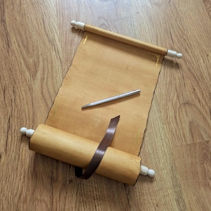 Pode incluir: Um pergaminho enrolado com pinos de madeira e pináculos brancos, uma caneta prateada e uma fita castanha sobre uma superfície de madeira. O pergaminho está desenrolado, revelando um papel em branco e envelhecido.