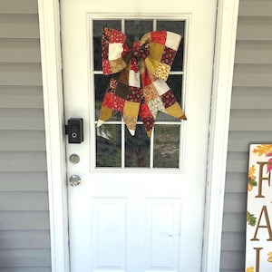 May include: A handmade, quilted bow in shades of red, gold, brown, and cream, hangs on a white door. The bow is made of various fabric squares, creating a patchwork design. The door has a glass window with a grid pattern.