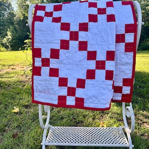 May include: A white and red patchwork quilt featuring a repeating block pattern and visible grid stitching. The quilt has a red border and the name June Ann embroidered in red cursive script on the lower left corner of the fabric surface.