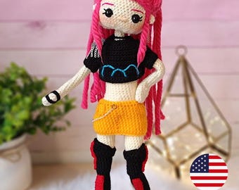 Puede incluir: Muñeca de ganchillo con pelo rosa, top negro, falda amarilla y botas negras con detalles rojos. La muñeca está etiquetada como "Huntrix girl K-pop". La muñeca está sobre una base de madera.
