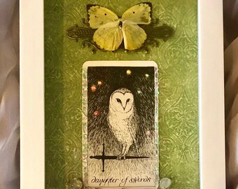 May include: Framed artwork with a yellow butterfly and a tarot card depicting an owl. The card is labelled "Daughter of Swords". The background is green with a swirling pattern, and the frame is white.