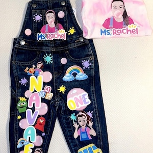 May include: Dark blue denim overalls and a pink tie-dye long-sleeve shirt. The overalls are decorated with colourful cartoon characters, the name "NAVAEA", and the number "ONE". The shirt and overalls feature the text "Ms. Rachel".