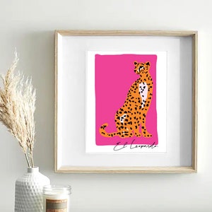 May include: A framed print of a cheetah sitting on a pink background. The cheetah is orange and black with white spots. The text "El Leopardo" is written below the cheetah.